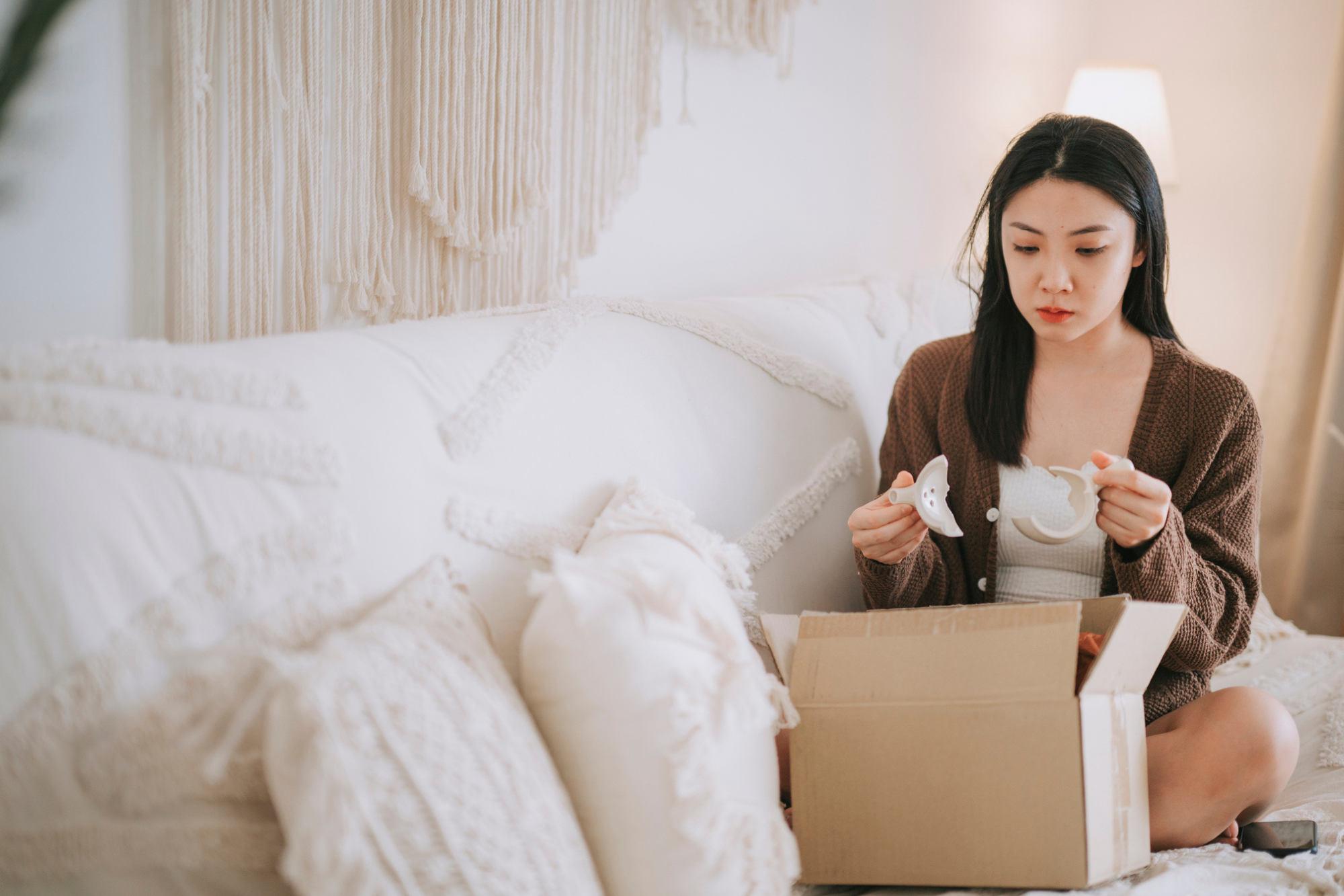 A female sitting on a couch with an open parcel holding a broken tea cup from her ecommerce order.